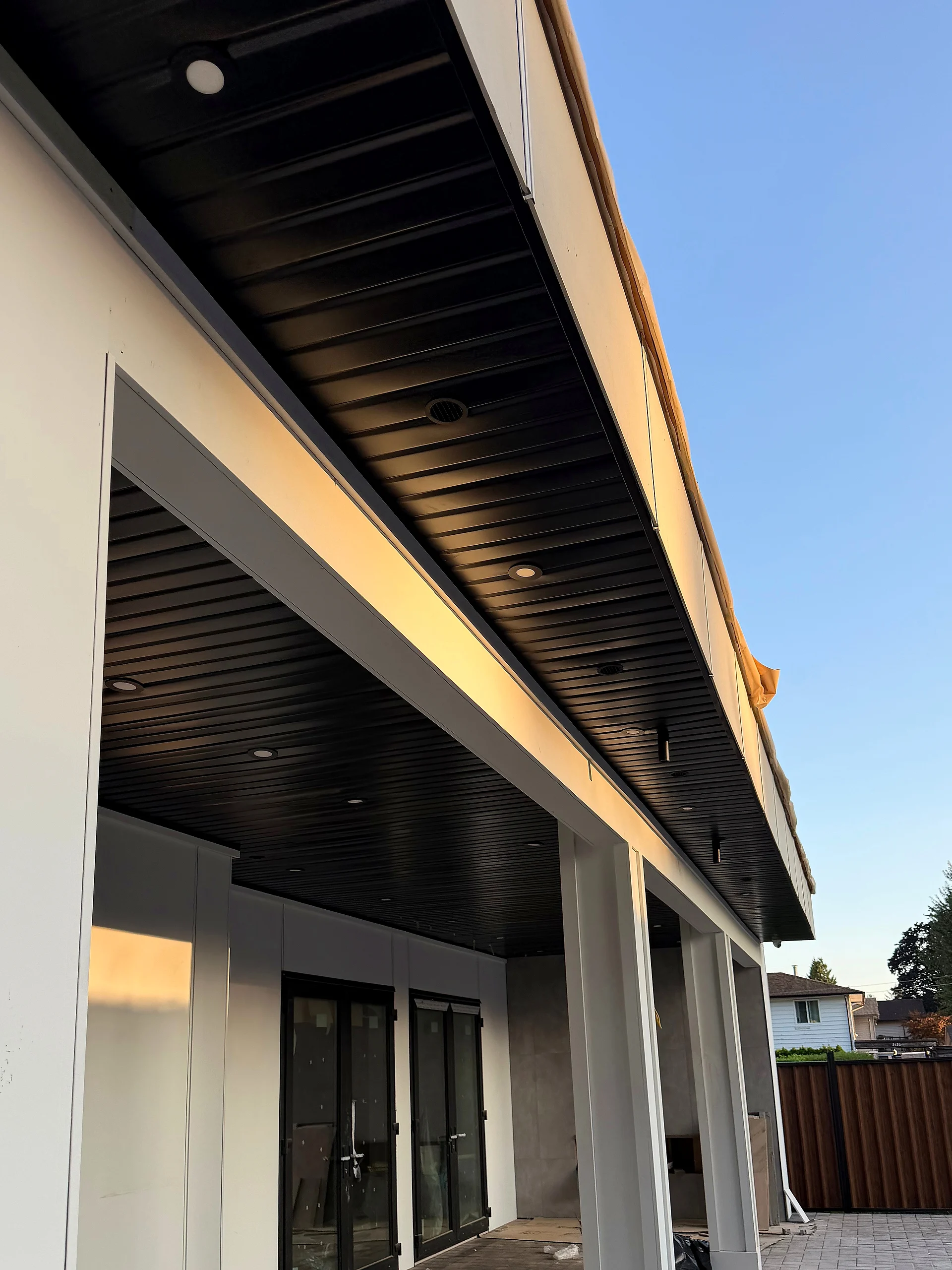 Covered patio with black ribbed soffit, white columns and golden evening light, Coquitlam BC