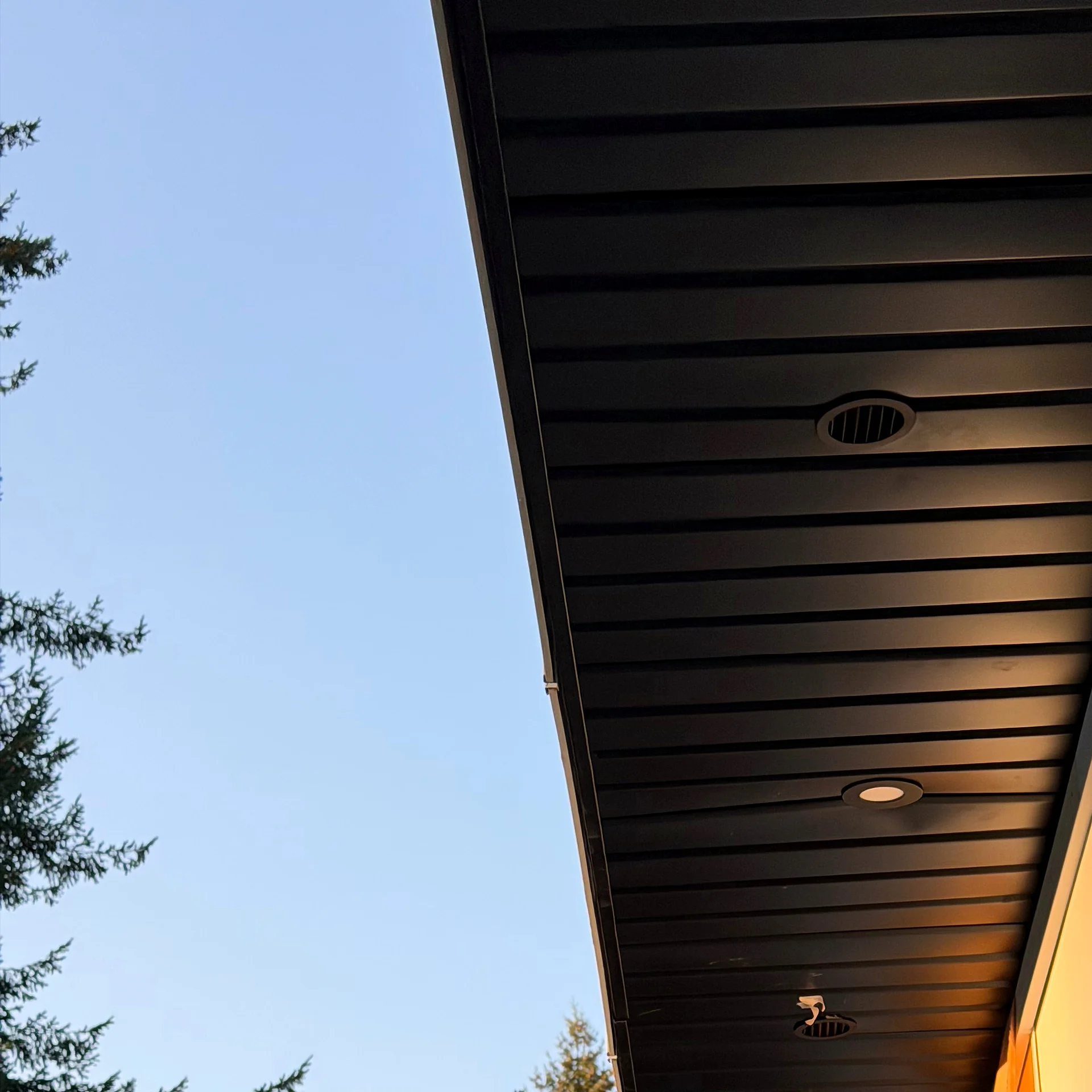 Black ribbed metal soffit along a side eave with a vent and recessed light in evening light, Coquitlam BC